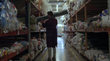 Bir adam (adam) bir fabrikada (bir depoda) bir iş ceket giyilen kumaş farklı rulo seçer. Konsept: İş, Depo, Tüketici, Handyman, Kalite Güvencesi.