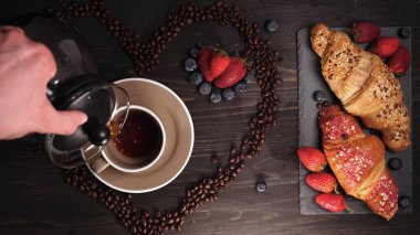 Ahşap bir masanın arka planında kahve çekirdeklerinin kalbi içinde bir kupa (kahve demleme) kahvesi buharda görülebilir. Tepside sırada kruvasan ve meyve var. Çilek ve yaban mersini etrafında.