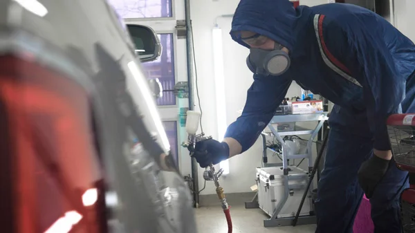 Professional in working uniform with spray guns in his hands and a respirator in protective glasses puts paint (ceramics) on the car on different parts of the car, flashlight with a folder in his hands checks a car engine for serviceability. Concept