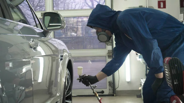 Professional in working uniform with spray guns in his hands and a respirator in protective glasses puts paint (ceramics) on the car on different parts of the car, flashlight with a folder in his hands checks a car engine for serviceability. Concept