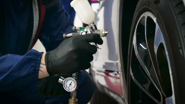 Professional in working uniform with spray guns in his hands and a respirator in protective glasses puts paint (ceramics) on the car on different parts of the car, flashlight with a folder in his hands checks a car engine for serviceability. Concept