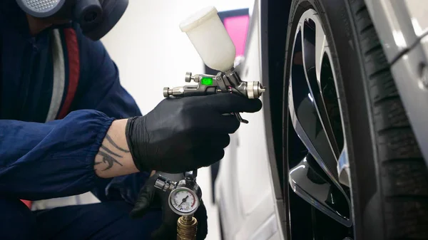 Professional in working uniform with spray guns in his hands and a respirator in protective glasses puts paint (ceramics) on the car on different parts of the car, flashlight with a folder in his hands checks a car engine for serviceability. Concept