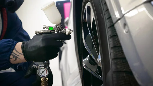 Professional in working uniform with spray guns in his hands and a respirator in protective glasses puts paint (ceramics) on the car on different parts of the car, flashlight with a folder in his hands checks a car engine for serviceability. Concept