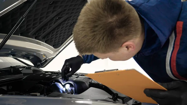 Professional in working uniform with spray guns in his hands and a respirator in protective glasses puts paint (ceramics) on the car on different parts of the car, flashlight with a folder in his hands checks a car engine for serviceability. Concept