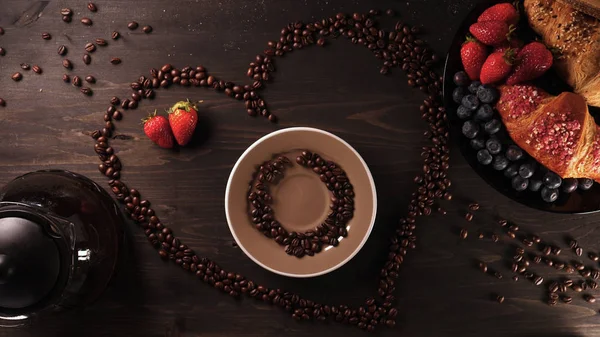 On the background of a wooden table the heart of coffee beans in it a mug (coffee brewing) coffee can be seen steam. Next on the tray are croissants and fruit. Strawberries and blueberries around.