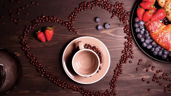 On the background of a wooden table the heart of coffee beans in it a mug (coffee brewing) coffee can be seen steam. Next on the tray are croissants and fruit. Strawberries and blueberries around.