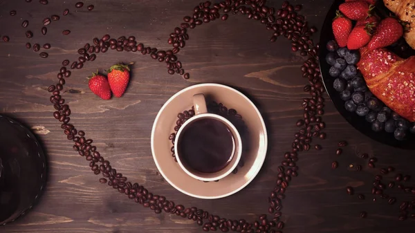 On the background of a wooden table the heart of coffee beans in it a mug (coffee brewing) coffee can be seen steam. Next on the tray are croissants and fruit. Strawberries and blueberries around.