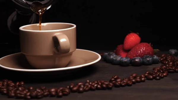 On the background of a wooden table the heart of coffee beans in it a mug (coffee brewing) coffee can be seen steam. Next on the tray are croissants and fruit. Strawberries and blueberries around.