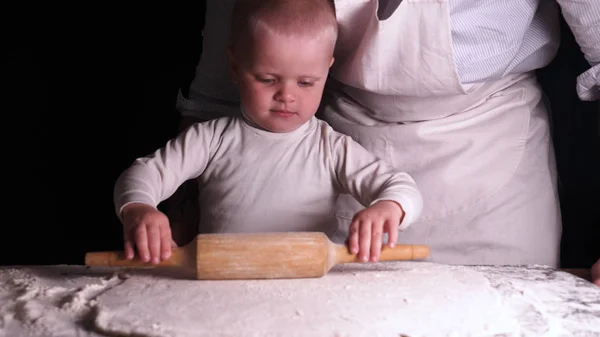 黒を背景にした小さな男の子は お父さん のシェフを助け ピザパンパンケーキやさまざまな小麦粉製品のためにロールアウトする生地を準備します コンセプト リトルシェフ ベイカー スローモーション — ストック写真