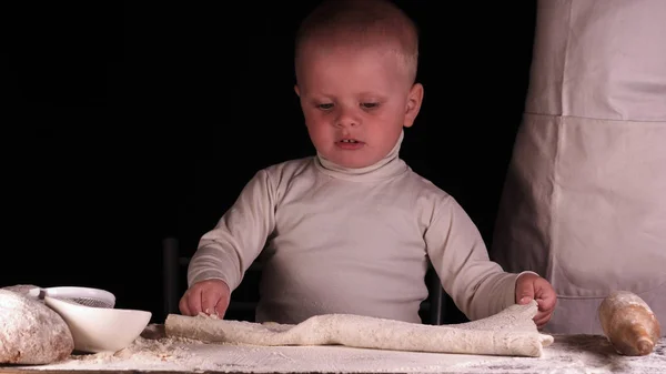 黒を背景にした小さな男の子は お父さん のシェフを助け ピザパンパンケーキやさまざまな小麦粉製品のためにロールアウトする生地を準備します コンセプト リトルシェフ ベイカー スローモーション — ストック写真
