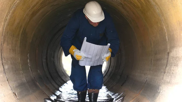 A worker in a working uniform, rubber boots checks the sewage canal inside it and write down on paper all that. Polluted water flows from the water channel. Concept of: Recording, Testing, Quality.