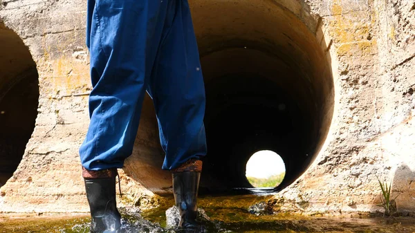 A worker in a working uniform, rubber boots checks the sewage canal inside it and write down on paper all that. Polluted water flows from the water channel. Concept of: Recording, Testing, Quality.