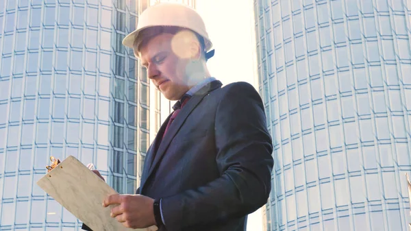 Builder or young handsome businessman, stands with his back looking at the buildings, wearing a white helmet and wearing a suit, talking on the phone, a skyscraper. Young handsome businessman talking on phone, in costume.