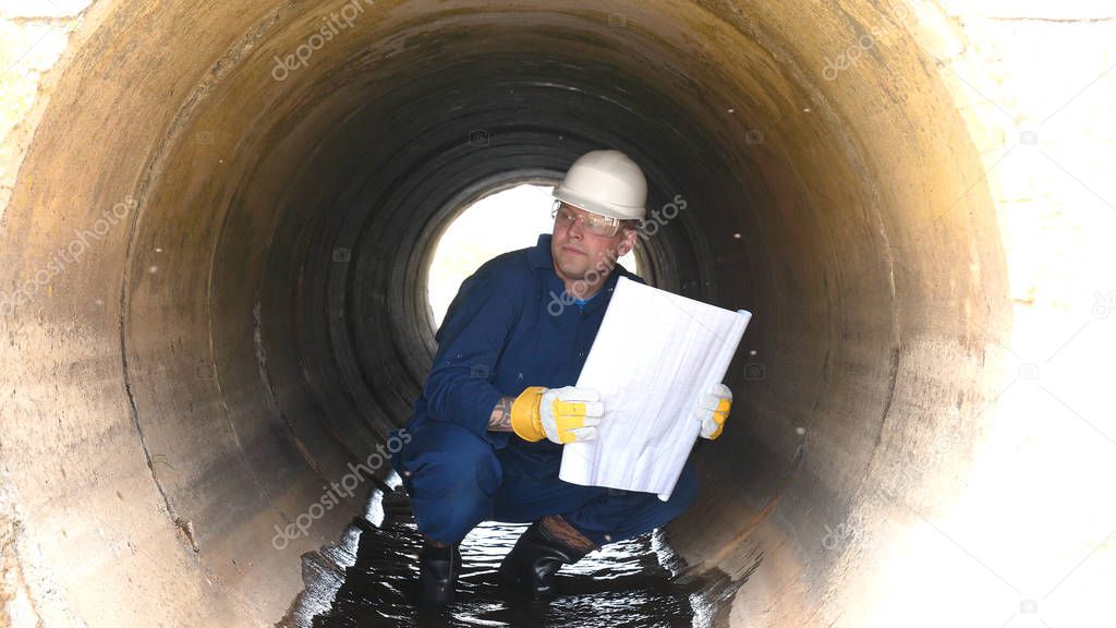 A worker in a working uniform, rubber boots checks the sewage canal inside it and write down on paper all that. Polluted water flows from the water channel. Concept of: Recording, Testing, Quality.