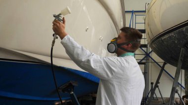 Young man (guy) specialist in white work coat polishes the yacht in the garage. Concept of: Preparation, Cleaning, Polishing, Master, Professional, After painting, Using chemistry. clipart