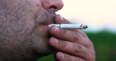 Doğanın arka planında, kıllı (sakallı) bir adam sigara içer, duman ıstırlar ve duman üfler. Kavramı: Sağlığa zarar, Sigara, Zarar prerode, Duman, El İşaretleme, Yavaş Mo.