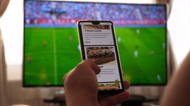 Evde TV arka plan (barda) karşı, adam (kadın) farklı atıştırmalıklar, pizza ve tatlılar cep telefonları kullanarak internet mağazaları üzerinden kaydırma sipariş. Konsept: futbol, spor, sipariş.