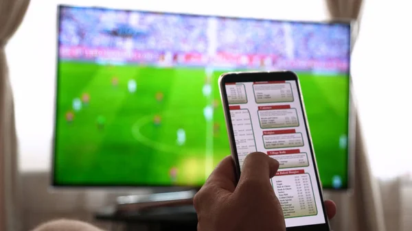 Evde TV arka plan (barda) karşı, adam (kadın) farklı atıştırmalıklar, pizza ve tatlılar cep telefonları kullanarak internet mağazaları üzerinden kaydırma sipariş. Konsept: futbol, spor, sipariş.