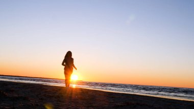 Günbatımının arka planında genç bir kadın deniz kıyısında güneş egzersizlerini koşarak, gerinerek ve düşerek yapar, denizdeki kız yoga yapar. Yavaş çekim, Günbatımı 'nın Portresi.