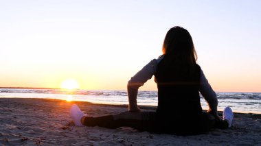 Günbatımının arka planında genç bir kadın deniz kıyısında güneş egzersizlerini koşarak, gerinerek ve düşerek yapar, denizdeki kız yoga yapar. Yavaş çekim, Günbatımı 'nın Portresi.