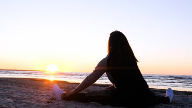 Günbatımının arka planında genç bir kadın deniz kıyısında güneş egzersizlerini koşarak, gerinerek ve düşerek yapar, denizdeki kız yoga yapar. Yavaş çekim, Günbatımı 'nın Portresi.