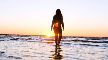 Günbatımının arka planında genç bir kadın deniz kıyısında güneş egzersizlerini koşarak, gerinerek ve düşerek yapar, denizdeki kız yoga yapar. Yavaş çekim, Günbatımı 'nın Portresi.