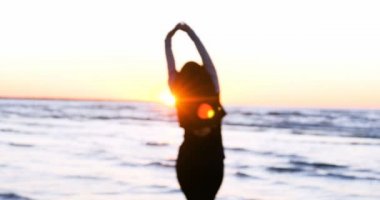 Günbatımının arka planında genç bir kadın deniz kıyısında güneş egzersizlerini koşarak, gerinerek ve düşerek yapar, denizdeki kız yoga yapar. Yavaş çekim, Günbatımı 'nın Portresi.