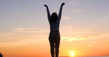 Günbatımının arka planında genç bir kadın deniz kıyısında güneş egzersizlerini koşarak, gerinerek ve düşerek yapar, denizdeki kız yoga yapar. Yavaş çekim, Günbatımı 'nın Portresi.