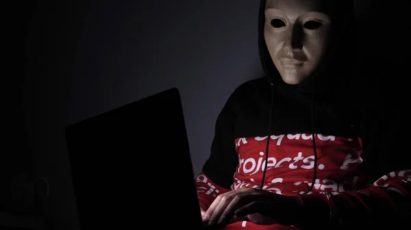Male hacker hidden face with the mask accessing to personal information on laptop (phone) in the dark. Technology, cyber crime concept.