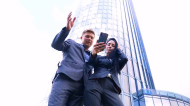 Gökdelen arka planında cep telefonuyla selfie çeken eğlenceli genç çift. Süit, İş, Çift, Mimarlık, Akıllı Telefon, Yaşam Tarzı, 5G Ağı.