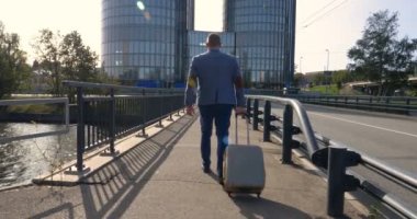 Seyahat eden işadamı tekerlekli bavuluyla şehrin silüetini geçiyor, iş adamı bavuluyla yürüyor ve cep telefonu kullanıyor..
