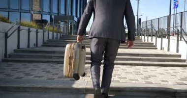 Seyahat eden işadamı tekerlekli bavuluyla şehrin silüetini geçiyor, iş adamı bavuluyla yürüyor ve cep telefonu kullanıyor..