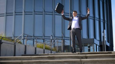 Başarıyı kutluyoruz. Heyecanlı genç işadamının kollarını havada tutması ve arka planda ofis binasıyla dışarıda durarak pozitifliğini ifade etmesi düşük açılı bir bakış açısı.