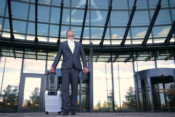 Traveling businessman walking with his wheeled suitcase across the city skyline, businessman walking with luggage and using mobile phone.