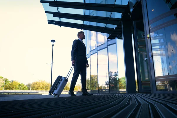 Traveling businessman walking with his wheeled suitcase across the city skyline, businessman walking with luggage and using mobile phone.