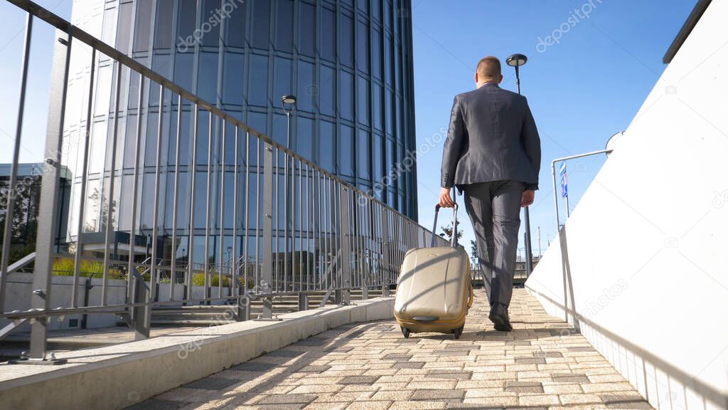 Traveling businessman walking with his wheeled suitcase across the city skyline, businessman walking with luggage and using mobile phone.