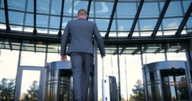 Seyahat eden işadamı tekerlekli bavuluyla şehrin silüetini geçiyor, iş adamı bavuluyla yürüyor ve cep telefonu kullanıyor..