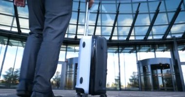 Seyahat eden işadamı tekerlekli bavuluyla şehrin silüetini geçiyor, iş adamı bavuluyla yürüyor ve cep telefonu kullanıyor..