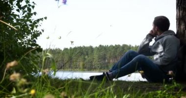 Adam yeşil çimlere oturur ve başka tarafa bakar. Ceketli, pantolonlu ve sırt çantalı genç adam nehre bakan bir kayanın üzerinde oturuyor..