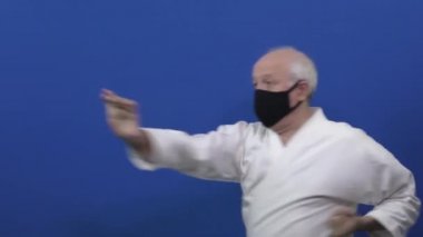 Siyah bir tıbbi maskeyle, bir atlet mavi arka planda duruşunu değiştirerek elleriyle vurur ve engeller. 