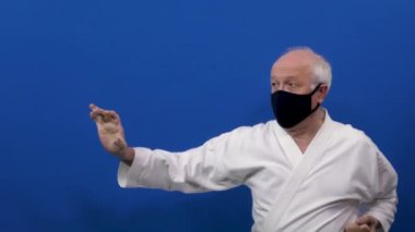 Siyah tıbbi maskeli mavi bir arka planda yaşlı bir erkek sporcu bloklar ve yumruklar eğitiyor.