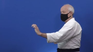 Siyah bir tıbbi maskeyle mavi bir arka planda yaşlı bir atlet kolları yumrukluyor ve blokluyor.