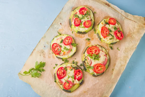 Healthy, nutritious zucchini small round pizza with pepperoni, tomatoes, cheese and parsley on parchment paper on light blue background.