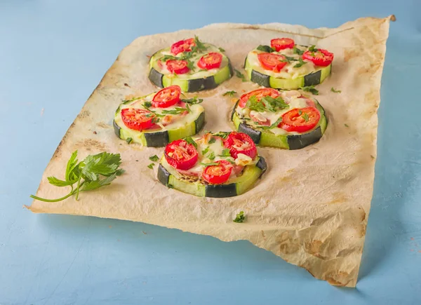 Healthy, nutritious zucchini small round pizza with pepperoni, tomatoes, cheese and parsley on parchment paper on light blue background.