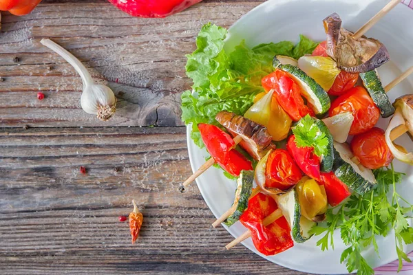 Healthy vegetable skewers. Organic fried vegetable shish kebab with eggplant, peppers, zucchini, tomatoes and onions on white plate with lettuce and parsley on wooden background.