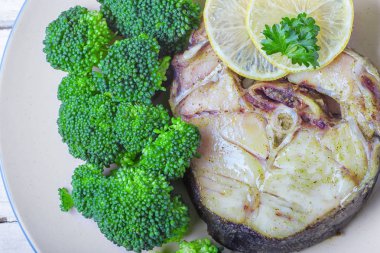 Vahşi siyah morina biftek brokoli, maydanoz ve limon ile hizmet verilen fırında pişirilir. Açık beyaz ahşap renkli, üstten görünüm.