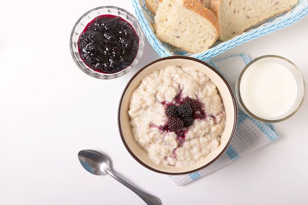 Healthy breakfast with a glass of milk, homemade oatmeal with blackberries in a blue bowl and fresh cereal bread on a light background.