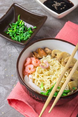 Deniz ürünleri ile Asya buğday erişte, mantar turşusu ve taze soğan, gri bir arka plan üzerinde bir kase susam serpilir. Asya tarzı, Japon yemekleri