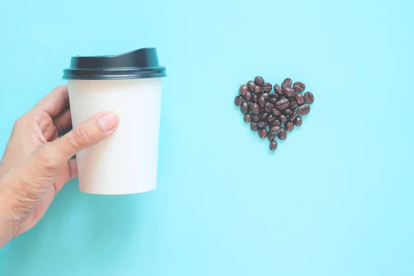 Donna mano tenendo tazzina da asporto caffè con chicchi di caffè a forma di cuore su sfondo color pastello. Pubblicità poster caffè — Foto Stock