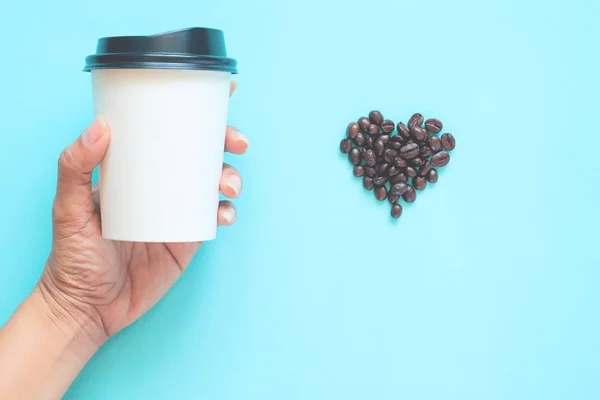 Donna mano tenendo tazzina da asporto caffè con chicchi di caffè a forma di cuore su sfondo color pastello — Foto Stock